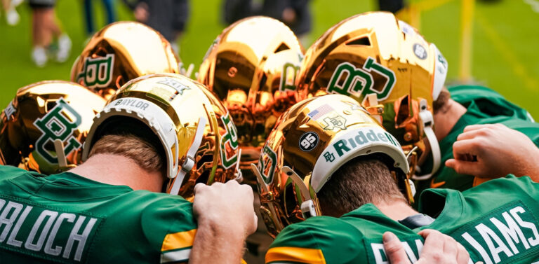 Baylor Bears football players huddle together on the field, heads down and arms around each other, wearing green jerseys and chrome gold helmets featuring the BU logo. Player names Bloch and Williams are visible on the jerseys.