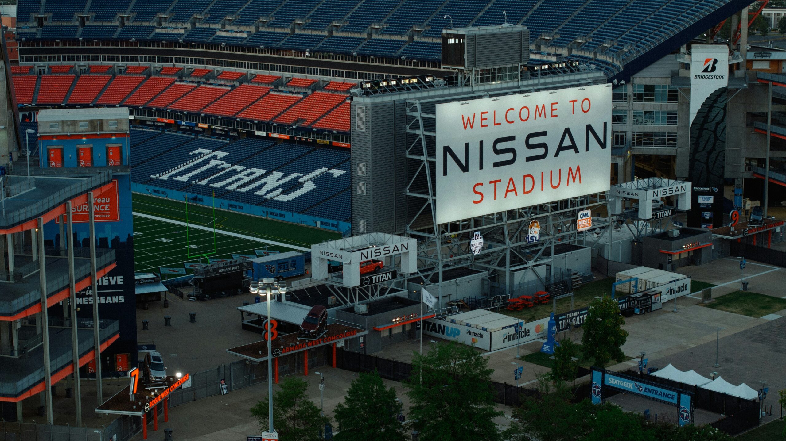 Aerial view of Nissan Stadium in Nashville, Tennessee, home of the NFL's Tennessee Titans, showing the field, blue and orange seating, and a large exterior welcome sign at dusk.