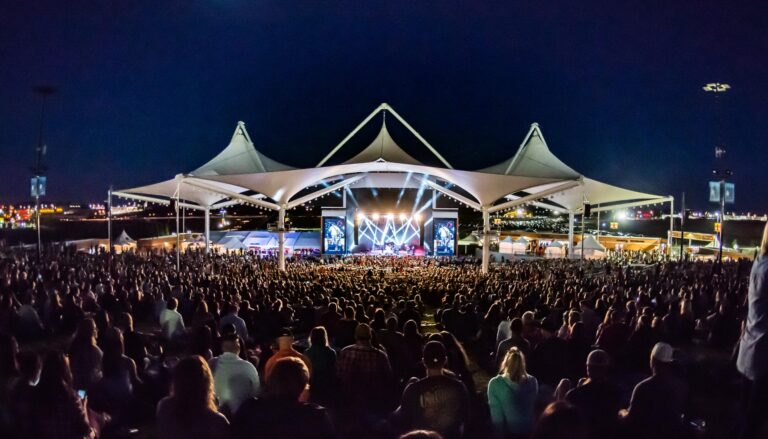 Arkansas Music Pavilion outdoor amphitheater at night, filled with a large crowd facing a lit stage beneath a white tensile fabric canopy structure with multiple peaked points. Large video screens flank the stage, illuminated by dramatic concert lighting against a dark blue sky.