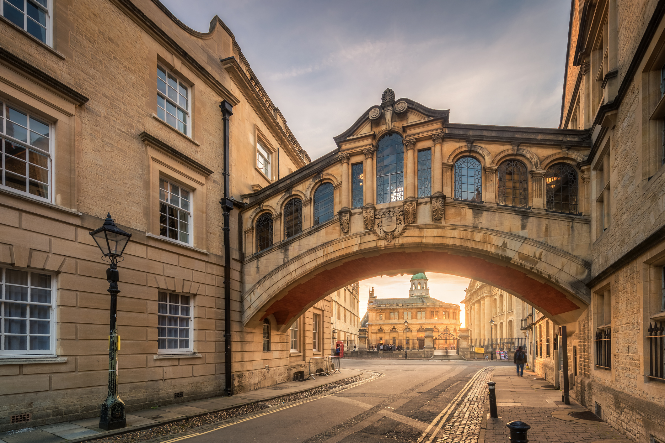 Oxford's Bridge of Sighs spanning a narrow lane at golden hour, with the Sheldonian Theatre visible through the arch and warm light illuminating the honey-colored stonework.