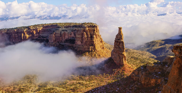 A tall sandstone spire rises from a fog-filled canyon surrounded by red rock cliffs and scrubby vegetation, with clouds rolling through the valley below a partly cloudy sky.