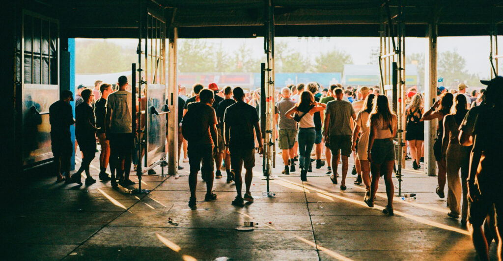 Festivalgoers stream through open gates into an outdoor music festival, backlit by warm golden sunlight, with a crowd and colorful event structures visible beyond the entrance.