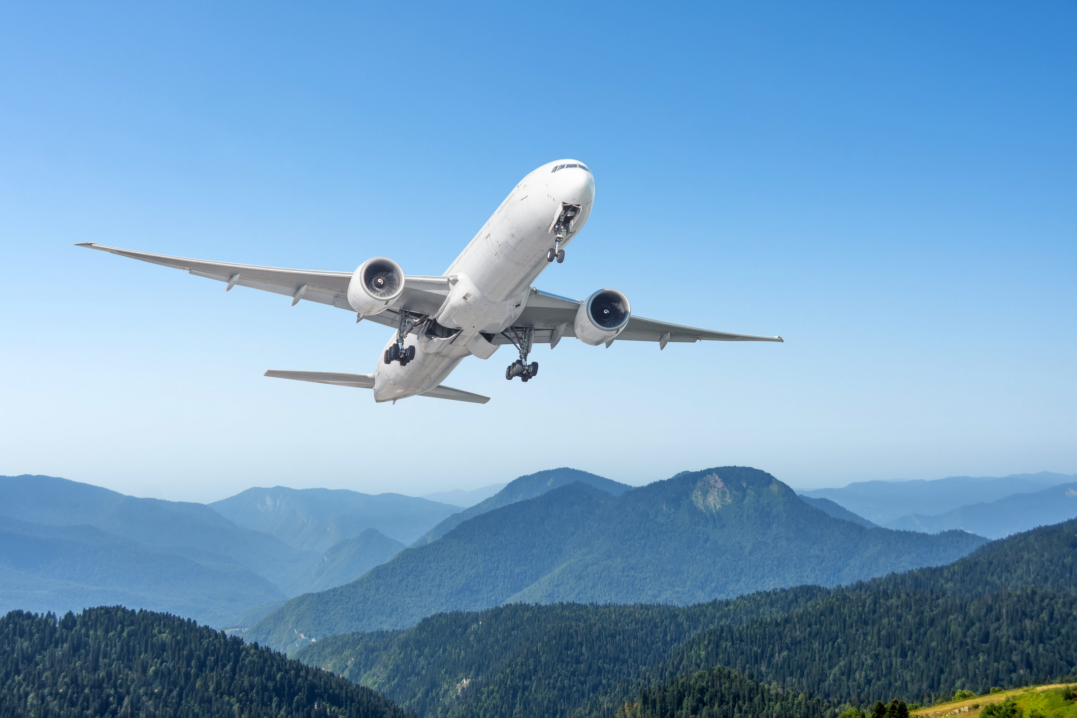 A white commercial aircraft climbs over a forested mountain range under a clear blue sky.