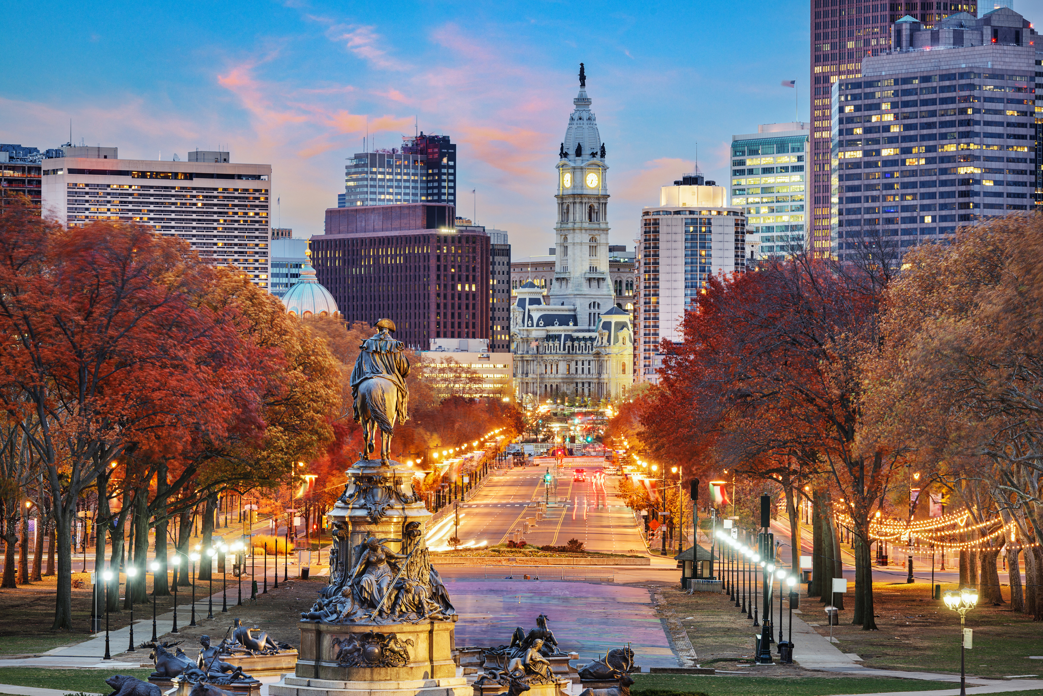 Philadelphia's Benjamin Franklin Parkway at dusk, looking toward City Hall with fall foliage, glowing streetlights, and the Washington monument statue in the foreground.