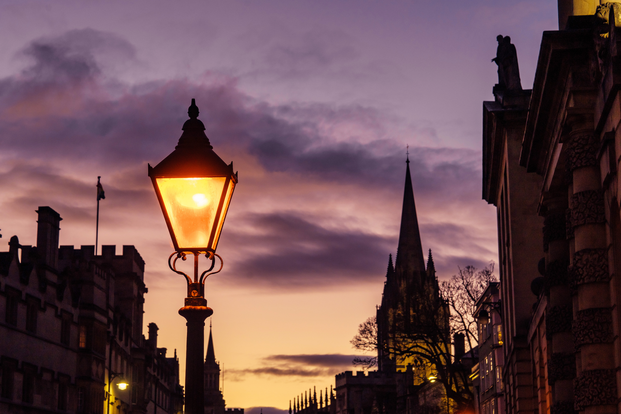 A glowing Victorian street lamp in the foreground of an Oxford city street at twilight, with a gothic church spire silhouetted against a purple sky.
