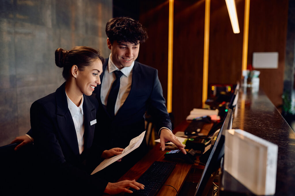 Two hotel front desk professionals in dark suits review information together at a computer monitor, with warm ambient lighting behind them.