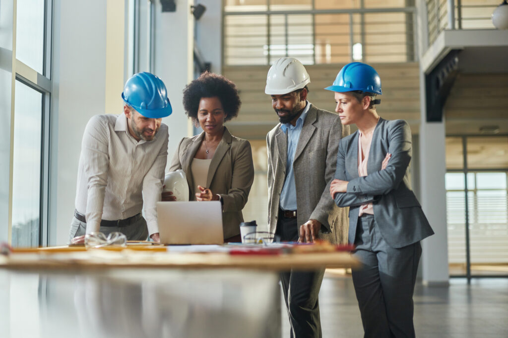 Four construction professionals in hard hats gather around a laptop on a worktable in a bright, modern building interior, reviewing project information together.