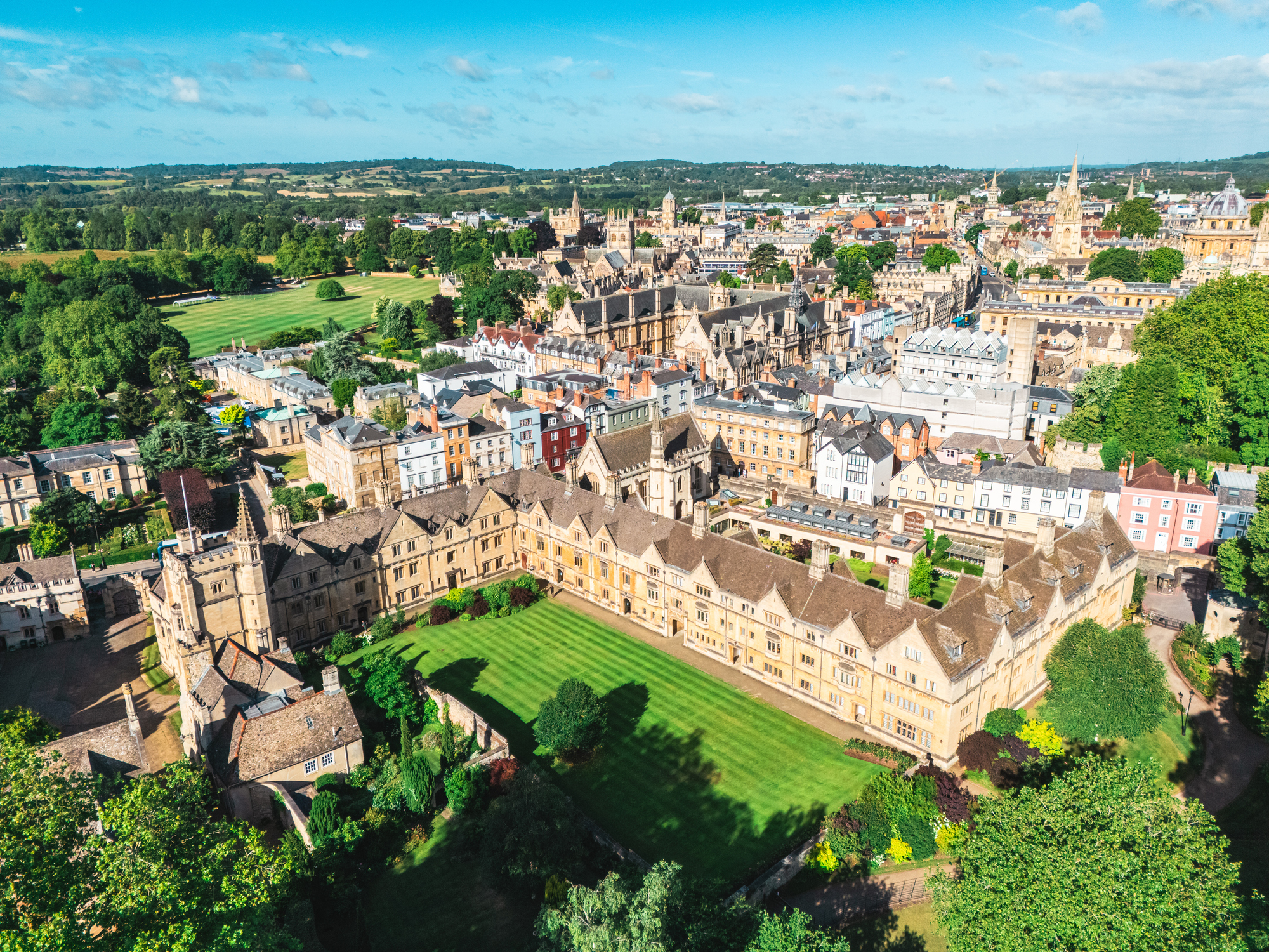 Aerial view of Oxford, England, showing the honey-stone college buildings, manicured quadrangle lawns, Gothic spires, and surrounding green countryside on a clear summer day.