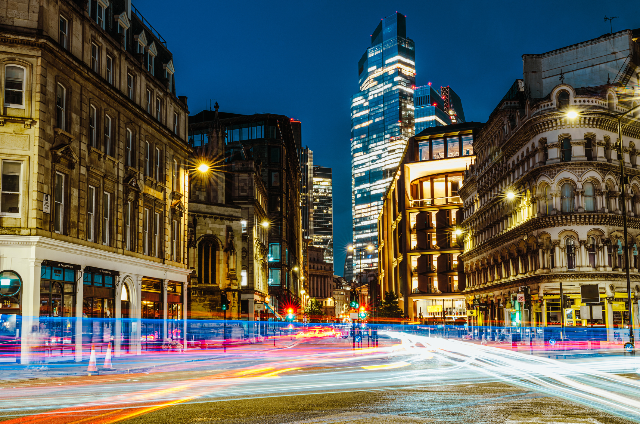 A long-exposure night shot of a city street with streaking vehicle light trails, historic stone buildings flanking the road, and a illuminated modern tower rising in the distance.