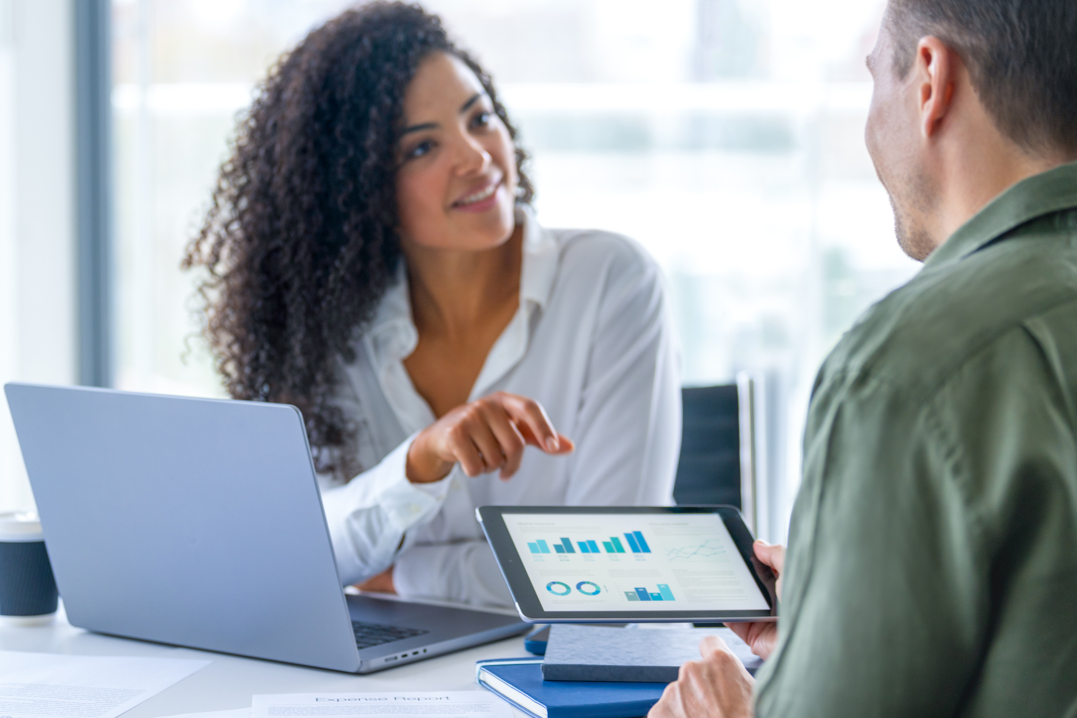 A woman points to data charts on a tablet held by a colleague during a meeting, with a laptop open on the table beside them in a bright office setting.