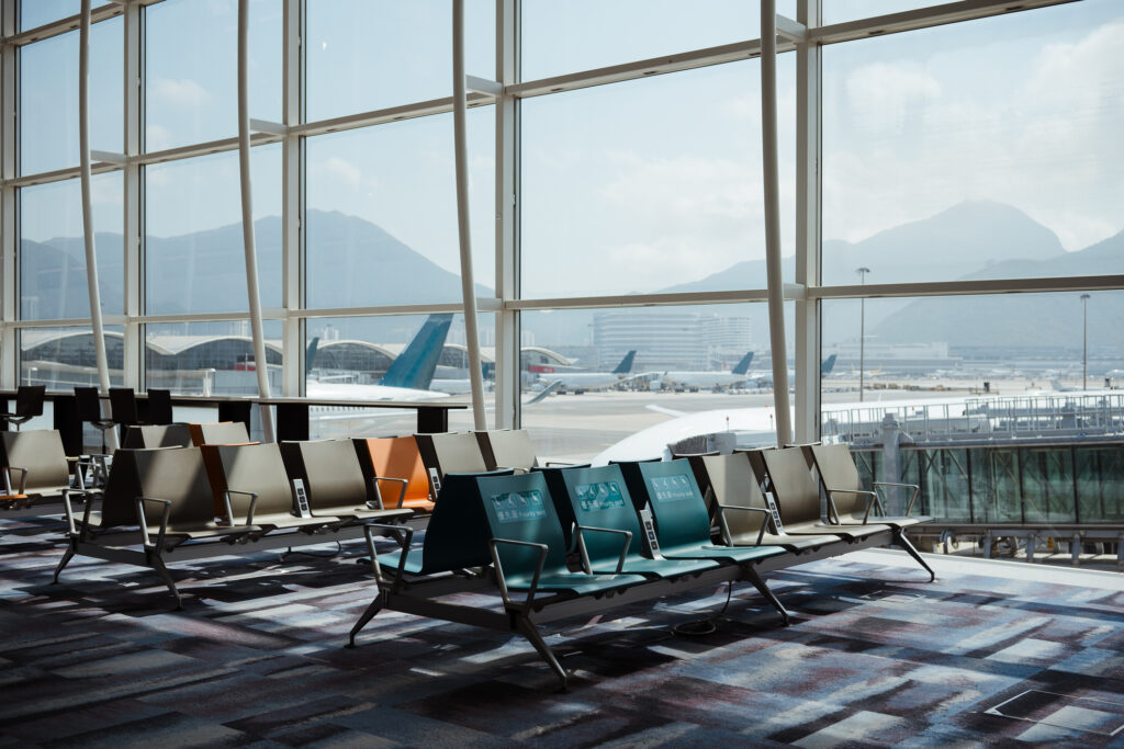 An empty airport gate lounge with rows of modern seating faces a floor-to-ceiling window overlooking a tarmac, parked aircraft, and mountains in the distance.