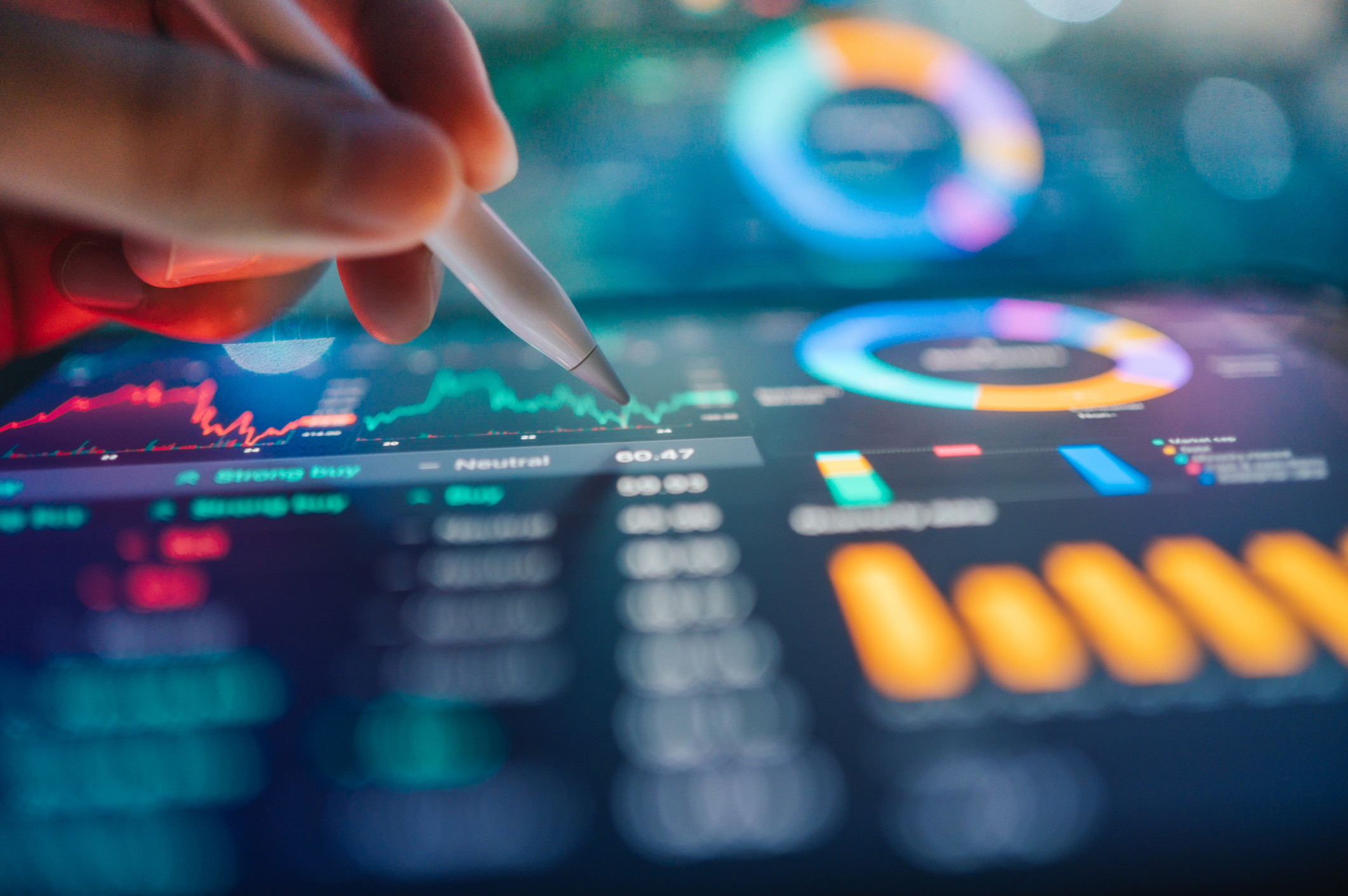 A hand using a stylus to interact with a tablet displaying a colorful financial analytics dashboard with line charts, donut charts, and bar graphs.