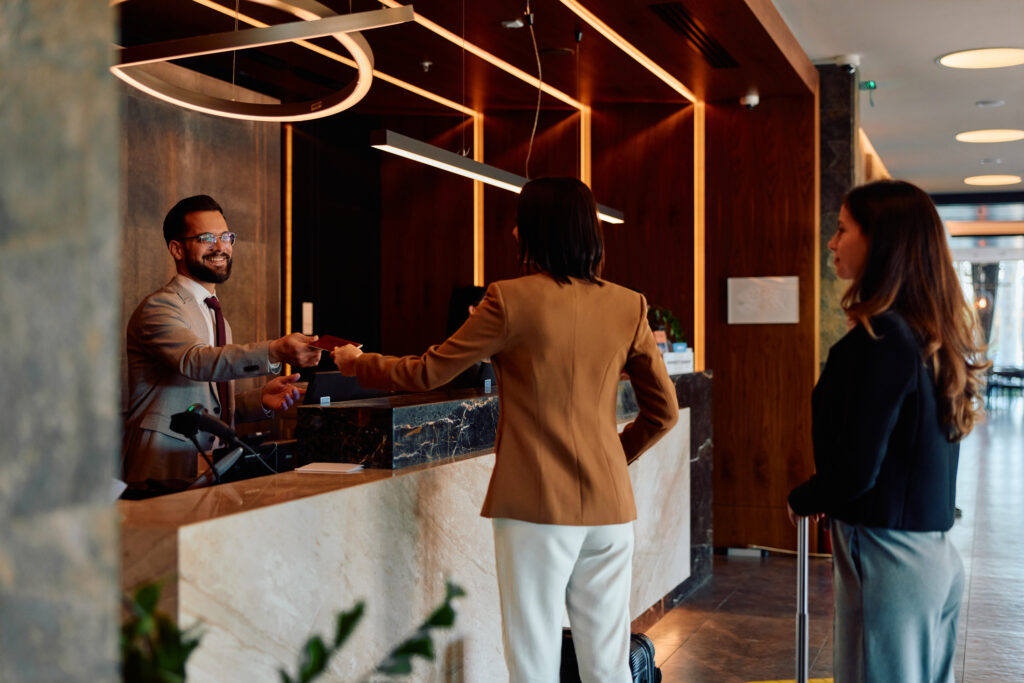 A smiling hotel front desk agent hands a room key card to a guest checking in, with a second guest waiting nearby in a warmly lit, upscale hotel lobby with marble and wood accents.