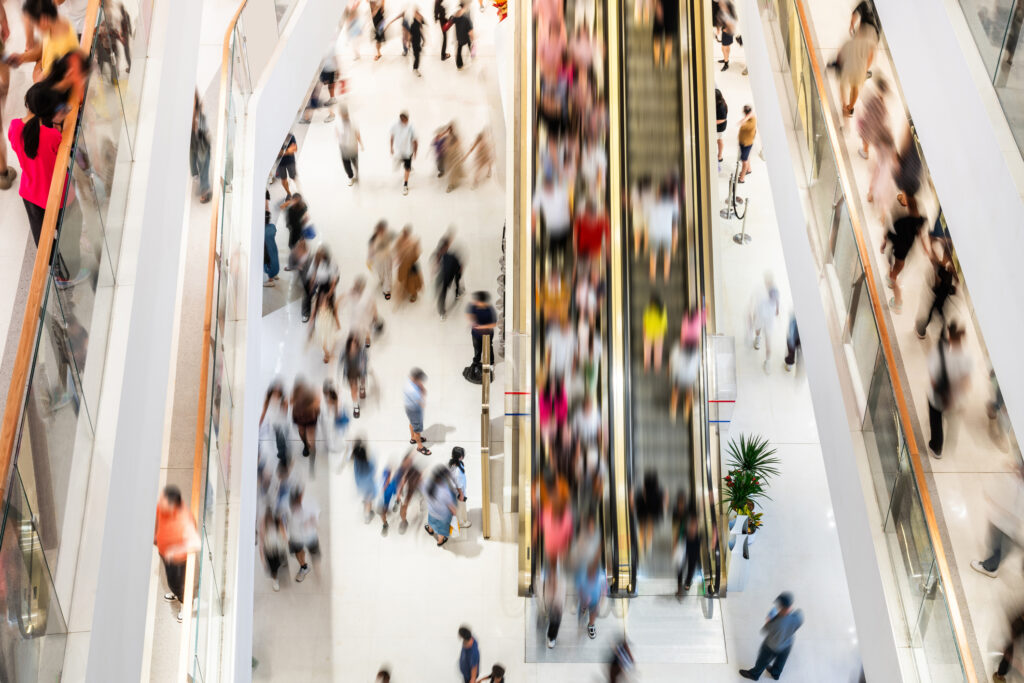 A long-exposure overhead shot of a busy shopping mall atrium, with crowds of shoppers blurred in motion across a bright white floor alongside a central escalator.