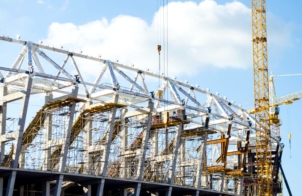 Steel roof trusses and scaffolding of a large stadium under construction, with a yellow tower crane visible against a blue sky.