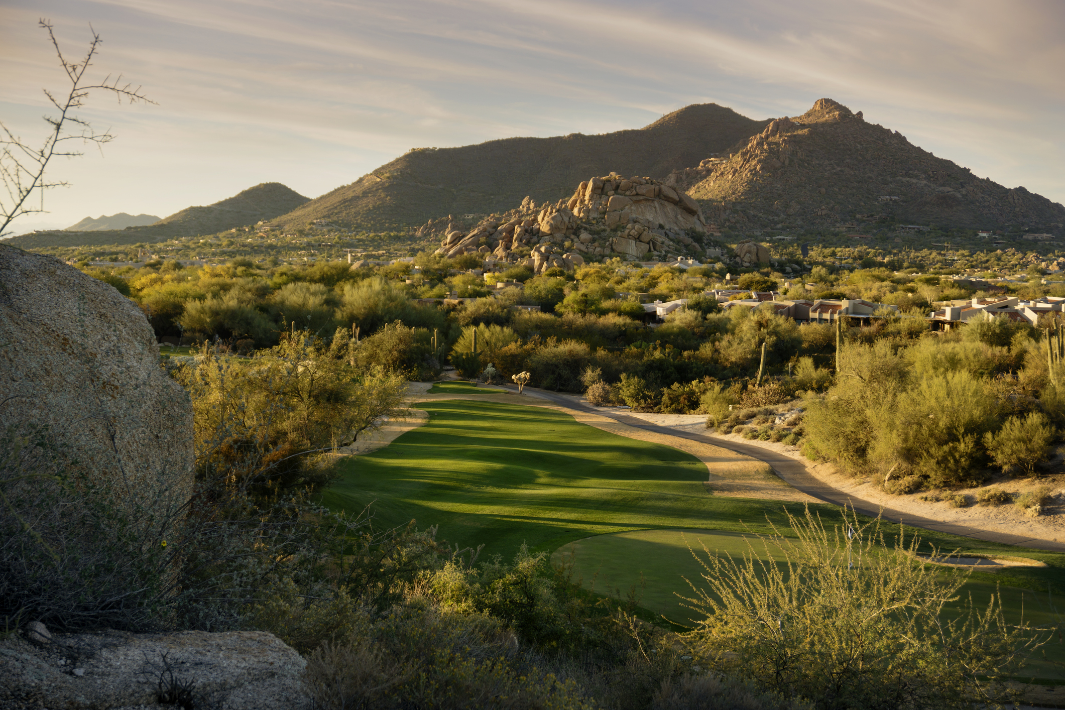 A desert golf course fairway winds through Sonoran desert vegetation with saguaro cacti and boulder formations, backed by rugged mountain peaks under a hazy sky.