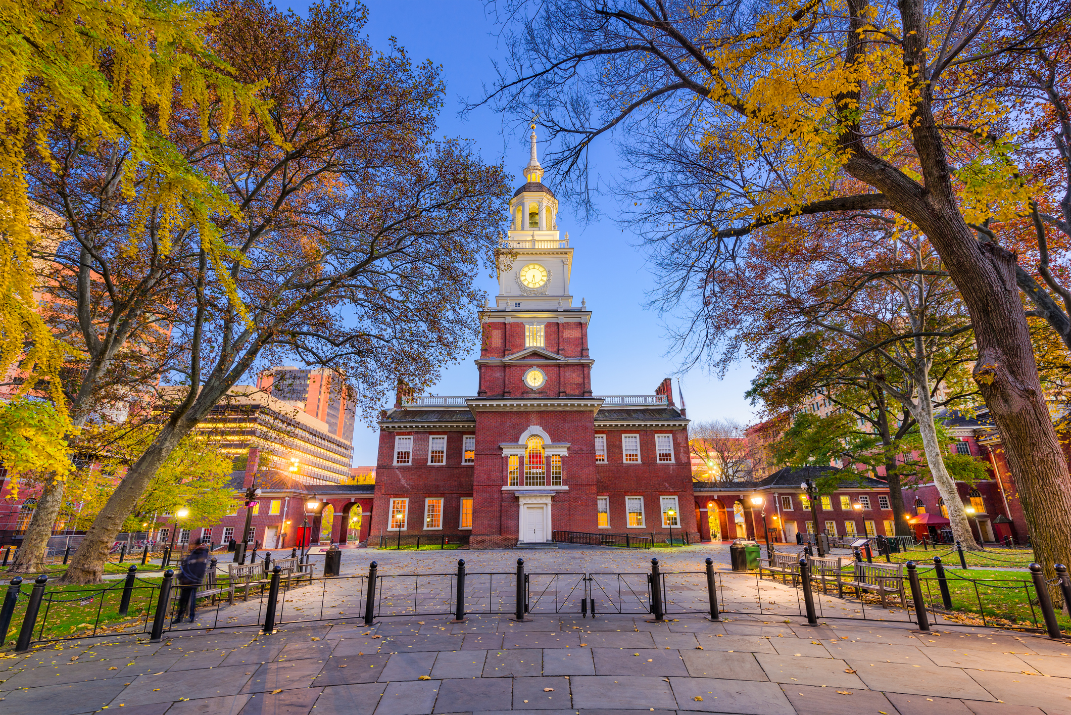 Independence Hall in Philadelphia at dusk, framed by autumn trees with its illuminated red brick facade and white clock tower against a blue sky.