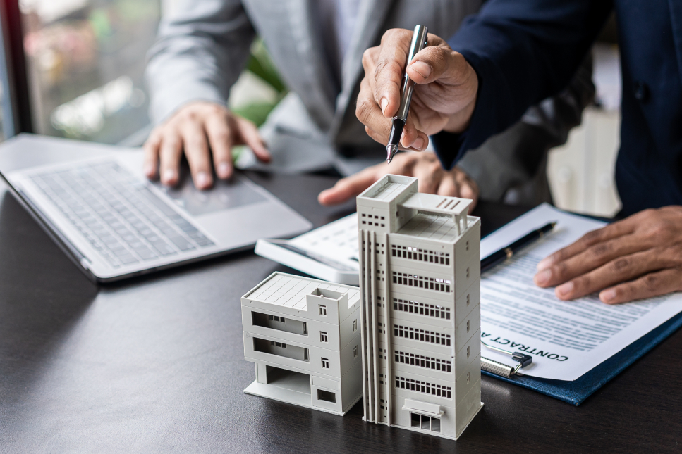 Two businesspeople review a real estate contract at a desk, with a small architectural building model, a laptop, and a contract document visible in the foreground.