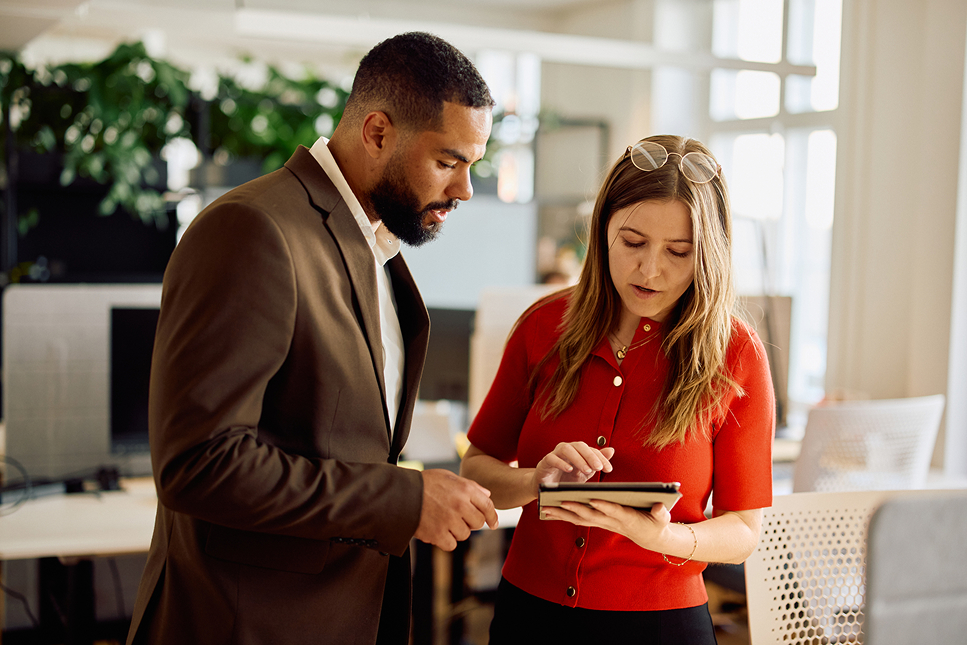Two colleagues review content on a tablet together in a bright, modern office. A man in a brown blazer and a woman in a red top stand side by side, both looking down at the screen she's holding.