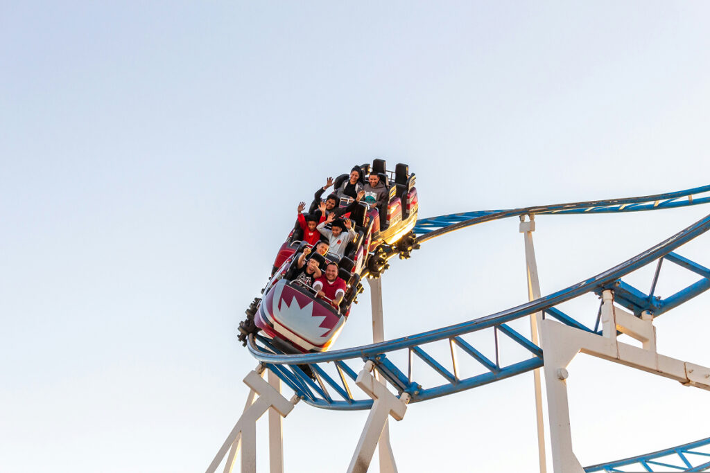 Riders wave and smile from a roller coaster car cresting a turn on a blue steel track against a clear sky.