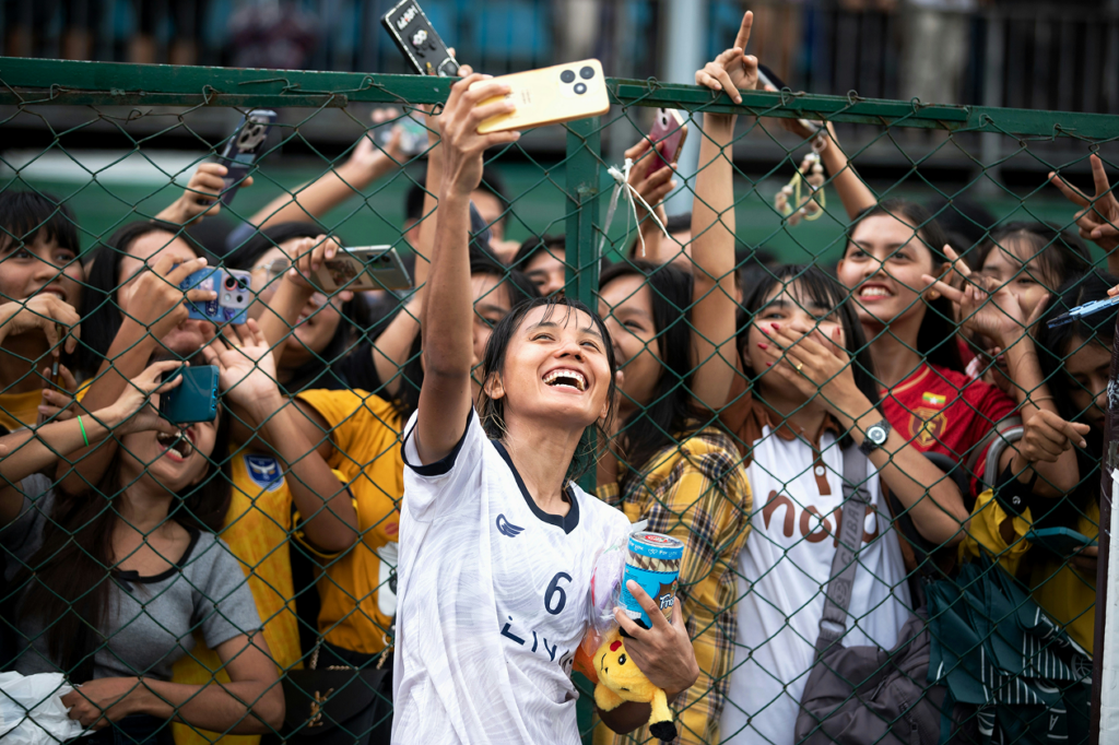 A soccer player wearing jersey number 6 laughs and takes a selfie against a chain-link fence as a crowd of fans reaches through to photograph her from the other side.