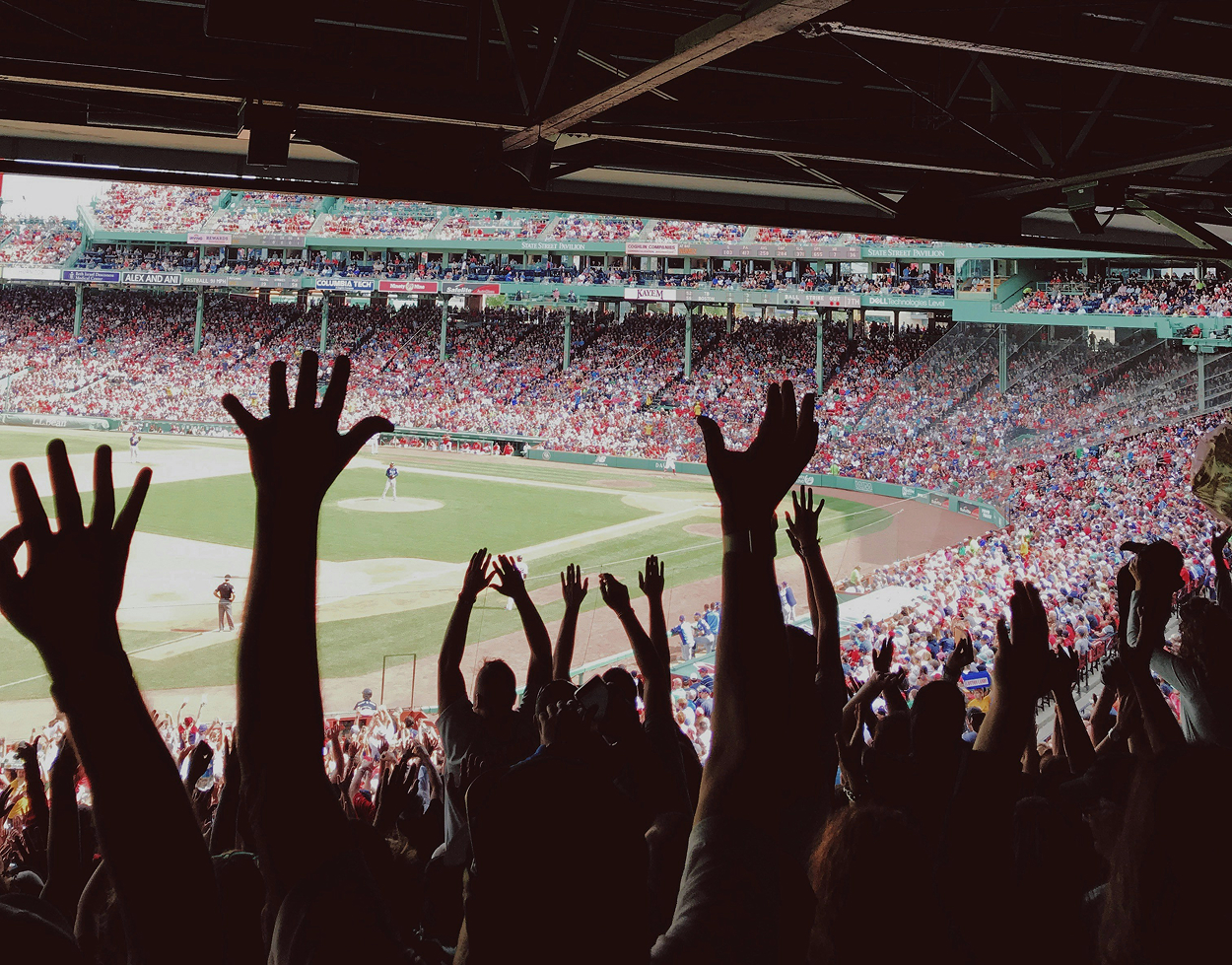 Silhouetted fans raise their hands in a packed baseball stadium, with the field and crowded grandstands visible in the background.