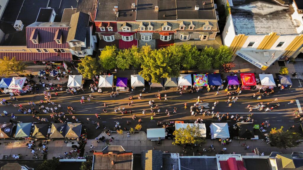Aerial view of a street festival in a historic urban neighborhood, with crowds strolling past colorful vendor tents lining a closed city block at golden hour.