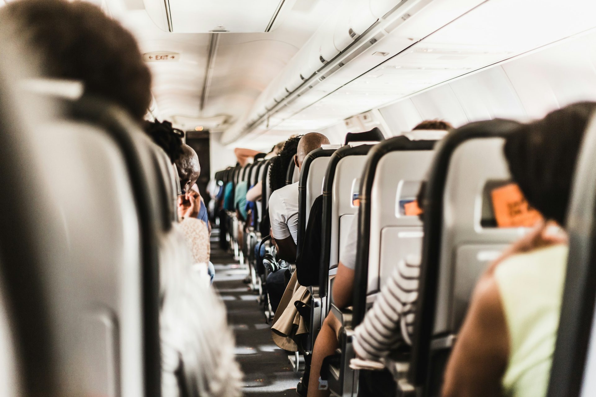 Passengers seated in the cabin of a full commercial aircraft, viewed from the aisle looking toward the front of the plane.