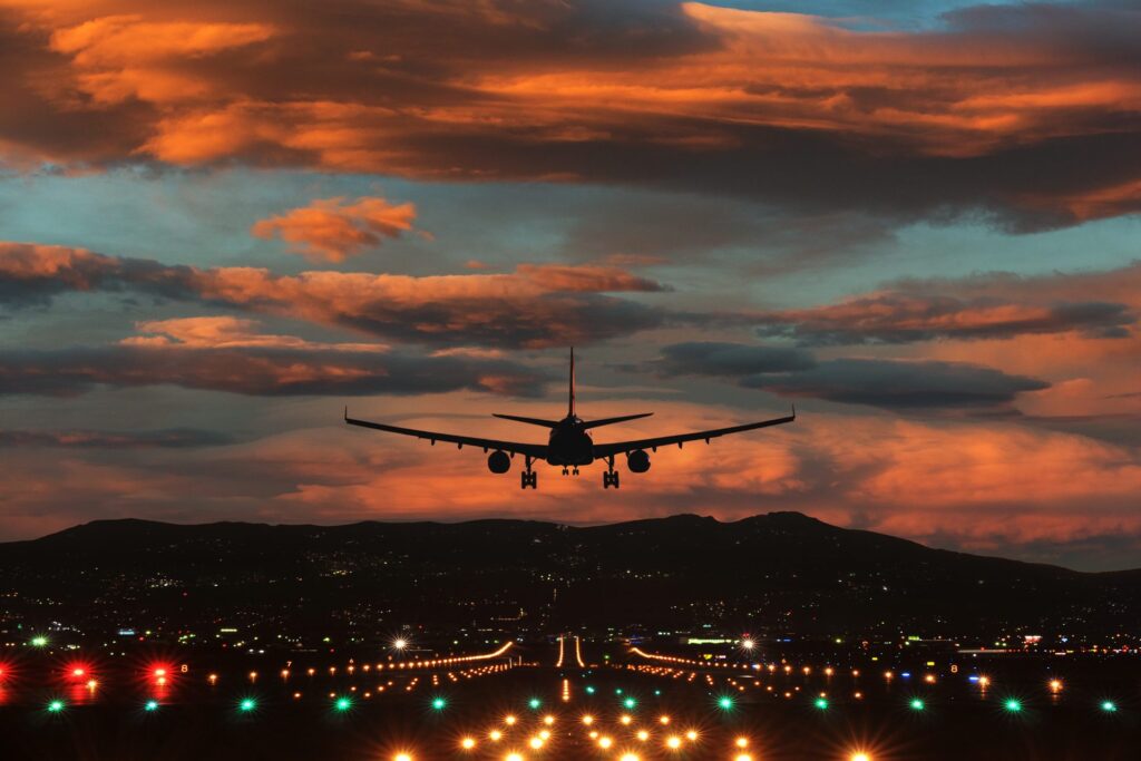 A commercial aircraft silhouetted against a dramatic sunset sky on final approach to an illuminated runway.