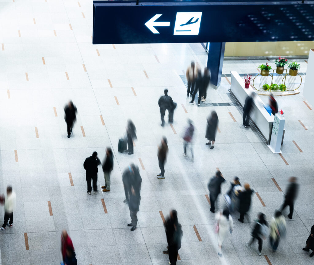 Overhead view of a busy airport terminal with motion-blurred travelers walking beneath a departures directional sign, conveying the energy of a bustling travel hub.