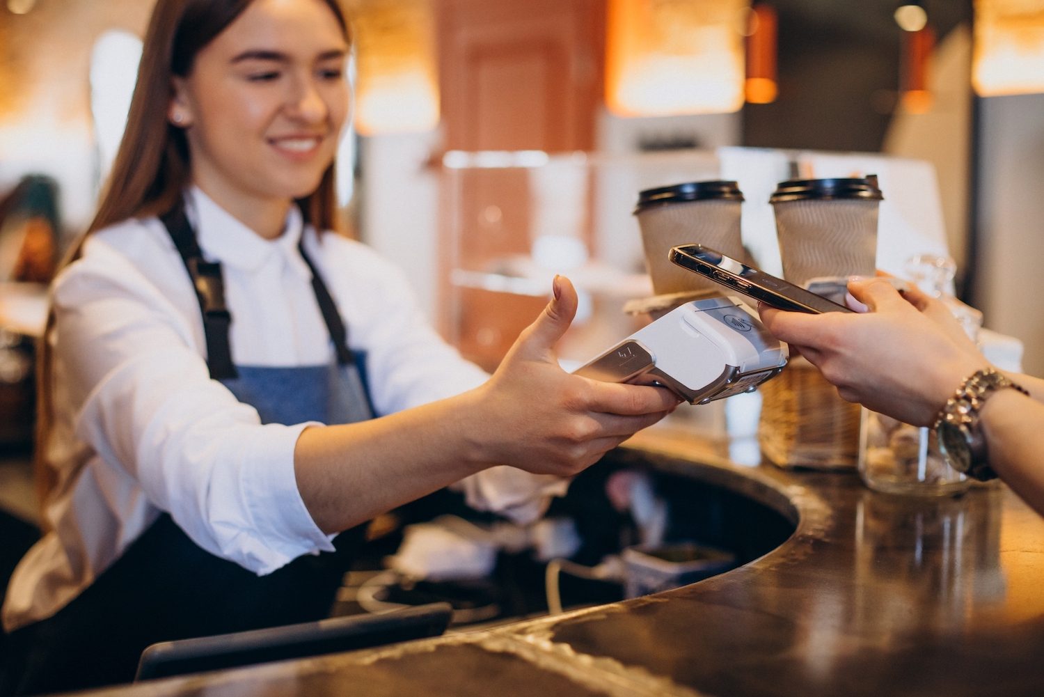 A smiling barista holds a card reader as a customer taps their phone to pay, with two to-go coffee cups on the counter in the background.