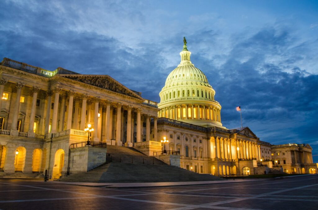 The U.S. Capitol building illuminated at dusk, with warm golden lighting on its neoclassical facade and dome set against a dramatic blue cloudy sky.