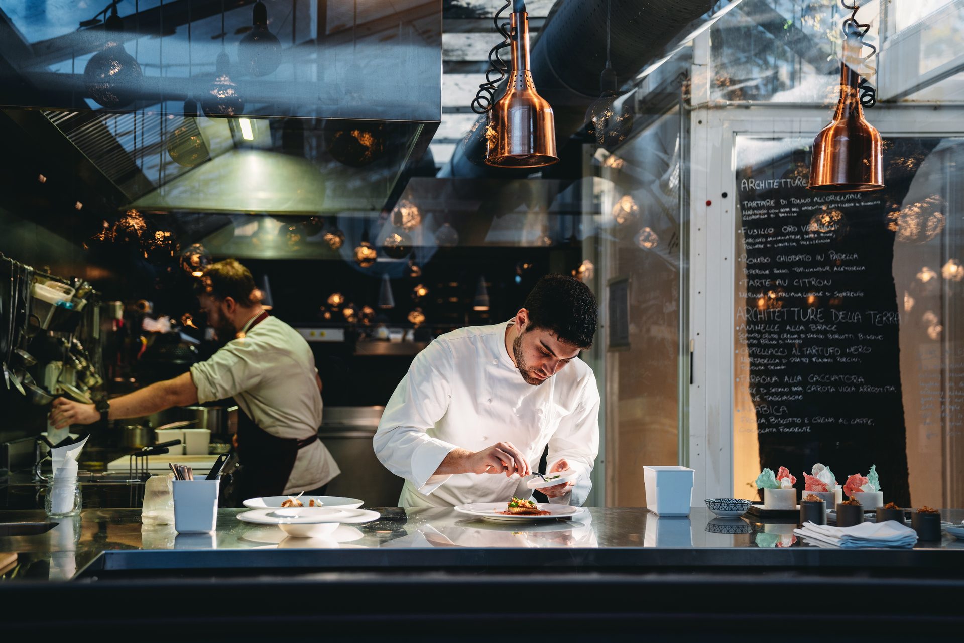 A chef in a white coat plates a dish at a stainless steel pass in an open kitchen, with copper heat lamps overhead and a handwritten chalkboard menu visible in the background.