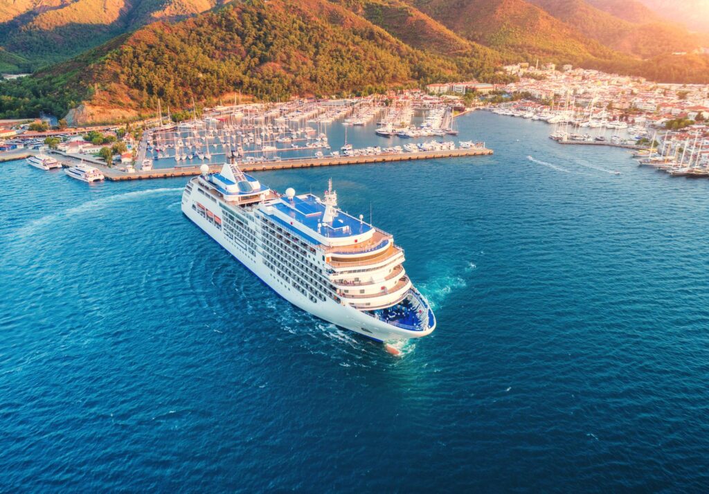 Aerial view of a large cruise ship departing a Mediterranean coastal town, with a marina full of sailboats, terracotta-roofed buildings, and forested hills in the background.
