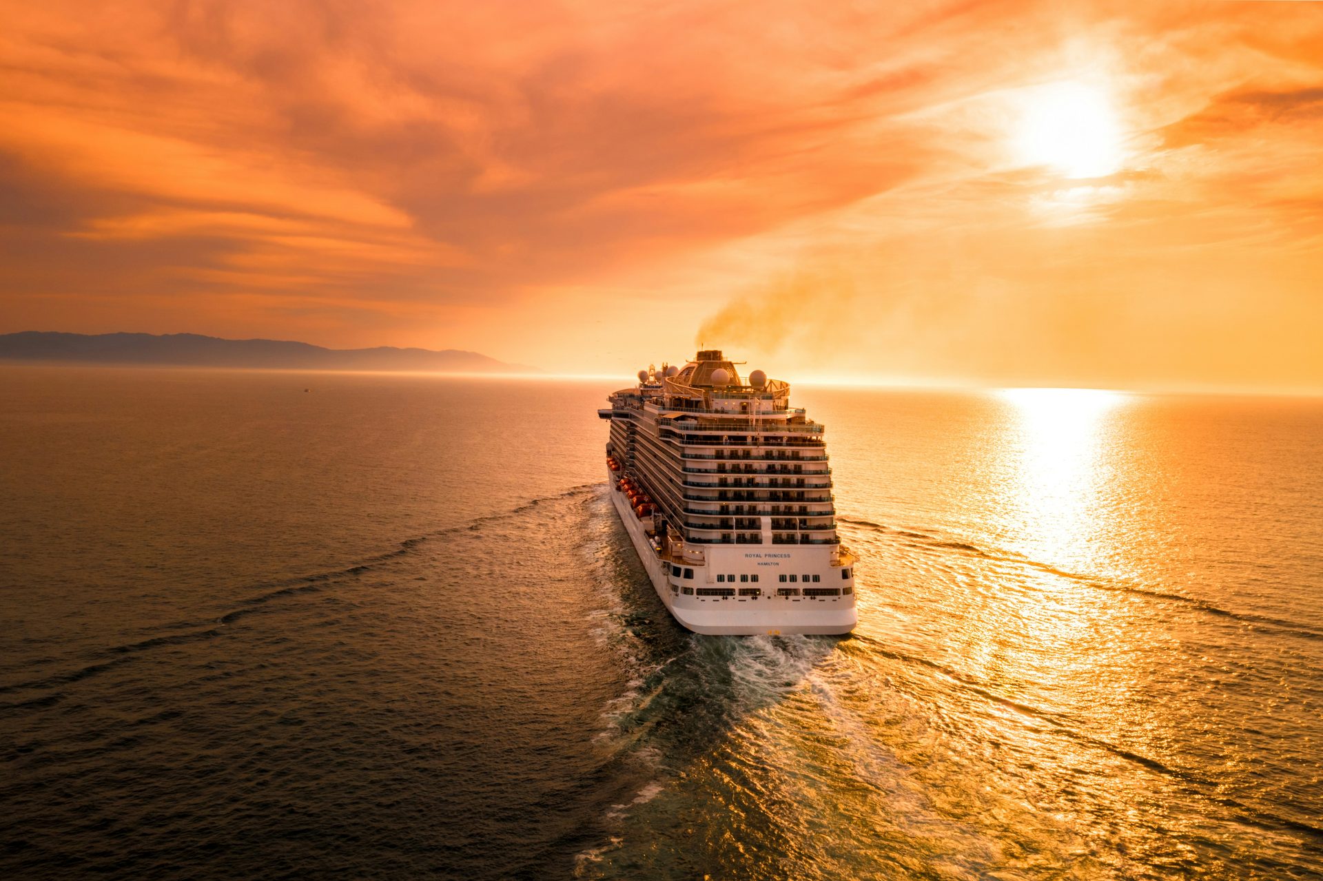 Aerial view of a large cruise ship sailing across open ocean at sunset, with a vivid orange and gold sky reflected on the water and distant mountains on the horizon.
