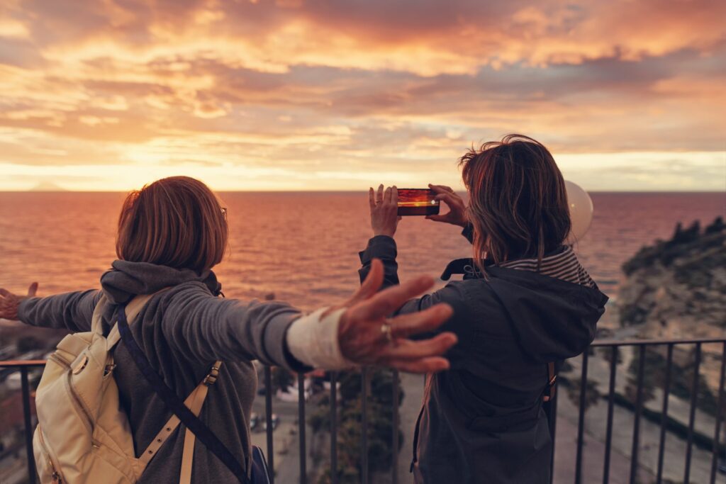 two female friends at a water overlook during sunset taking a photo with a smartphone
