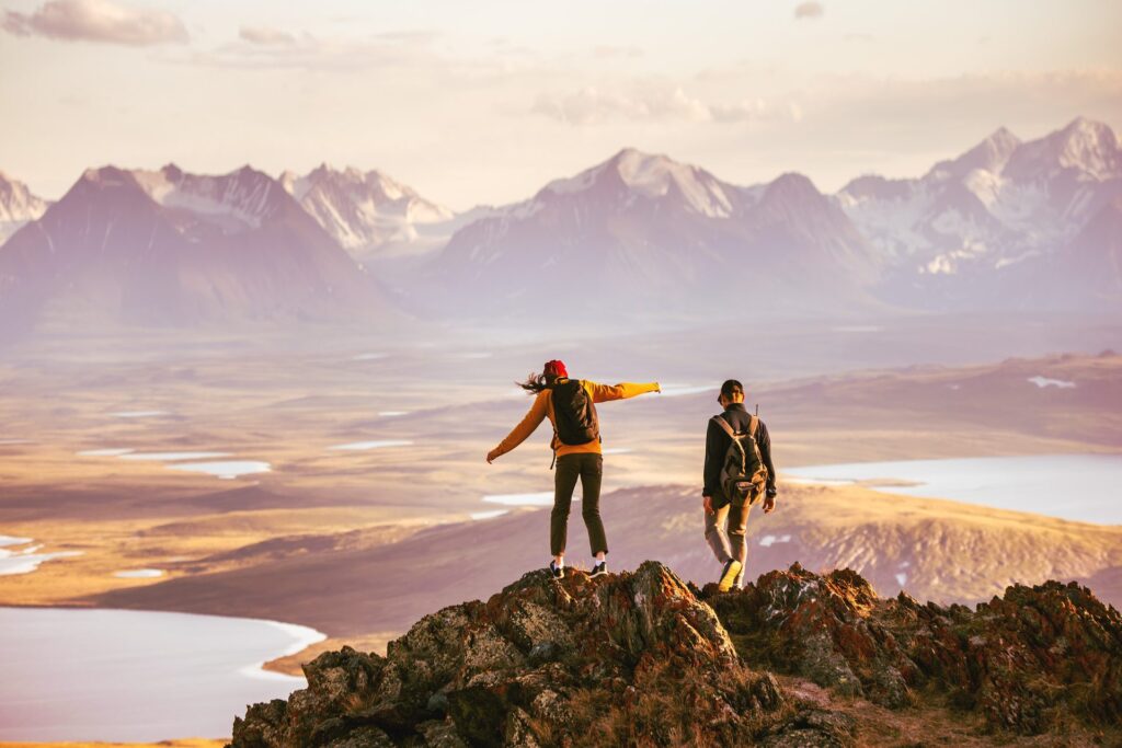 Two hikers with backpacks stand on a rocky mountain summit, one with arms outstretched, overlooking a vast landscape of lakes, valleys, and snow-capped peaks at golden hour.