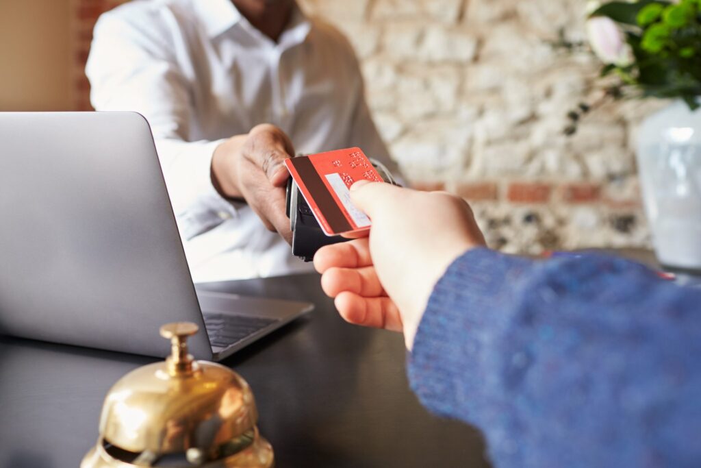 A guest hands a credit card to a hotel front desk agent at check-in, with a brass service bell and laptop visible on the counter.