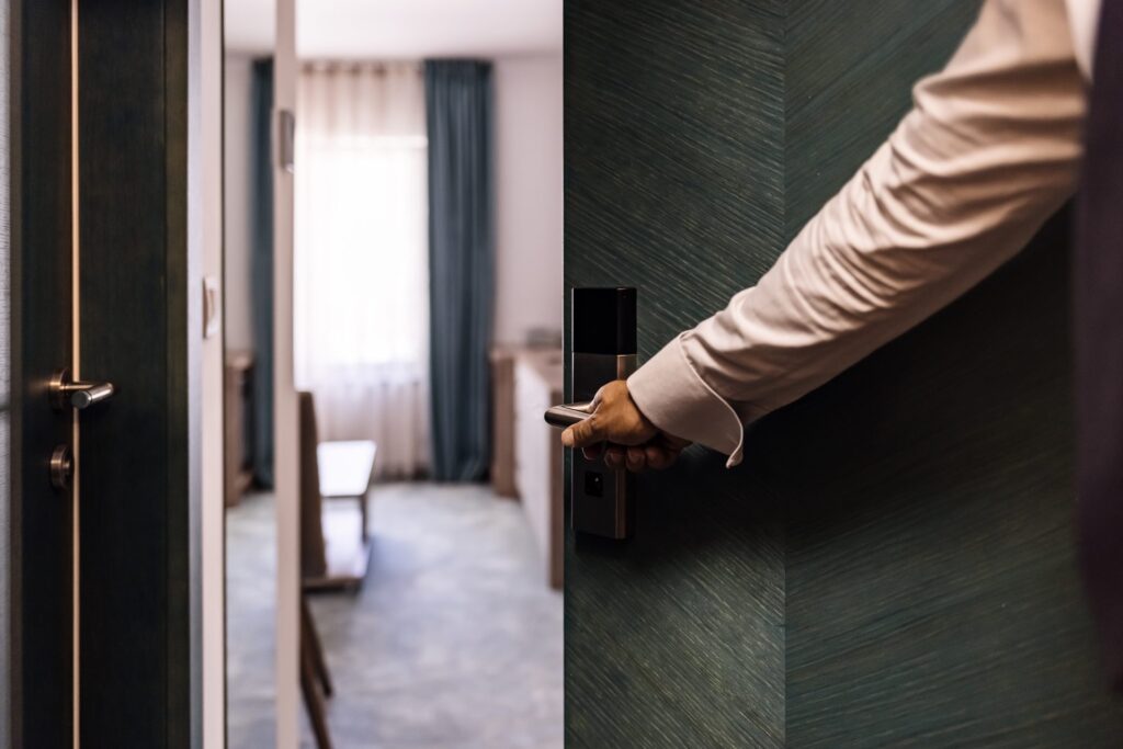 A hotel guest uses a key card to unlock a room door, with a neatly furnished room visible in the background.