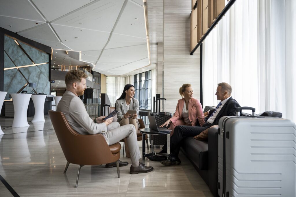Four business travelers converse in a modern hotel or airport lounge, seated in a circle with luggage nearby, in a bright, upscale interior with floor-to-ceiling windows.