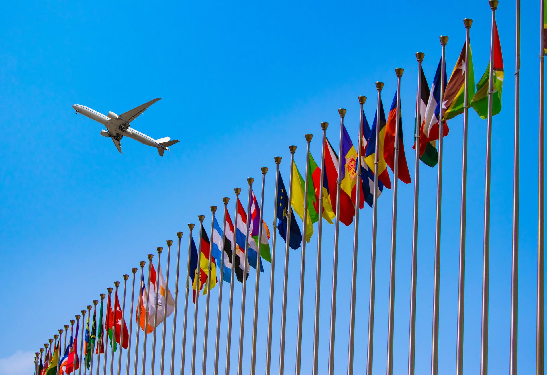 A commercial airplane flies over a curved row of international flags from nations around the world, against a bright blue sky, symbolizing global travel and international relations.