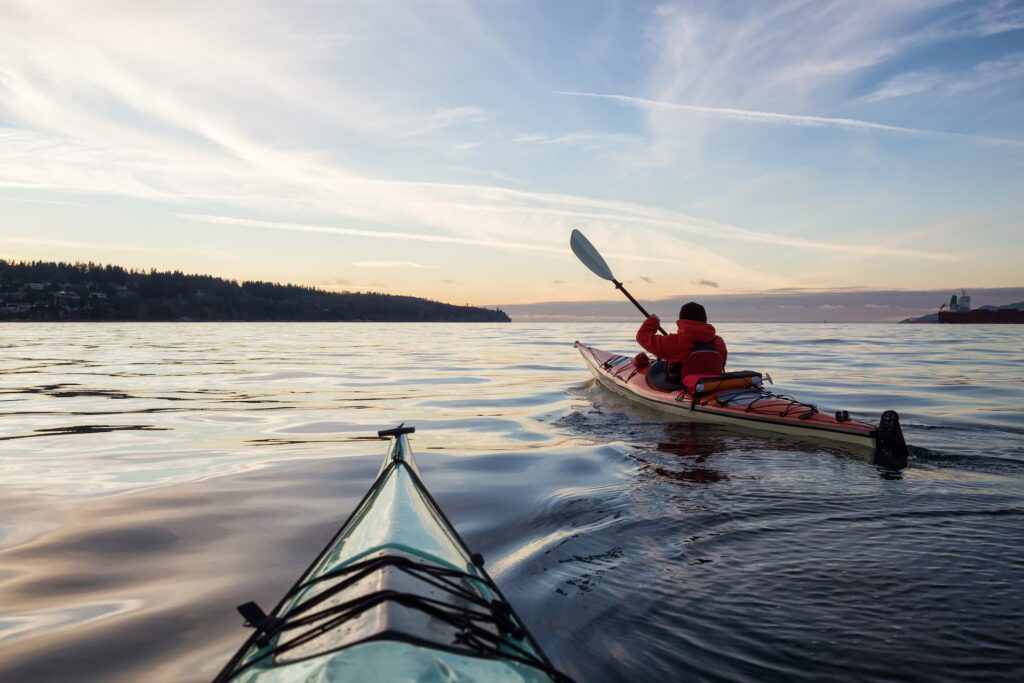 Two kayakers paddle across calm open water at dusk, viewed from the bow of one kayak looking toward a second paddler in an orange jacket, with a forested shoreline and soft sunset sky in the distance.
