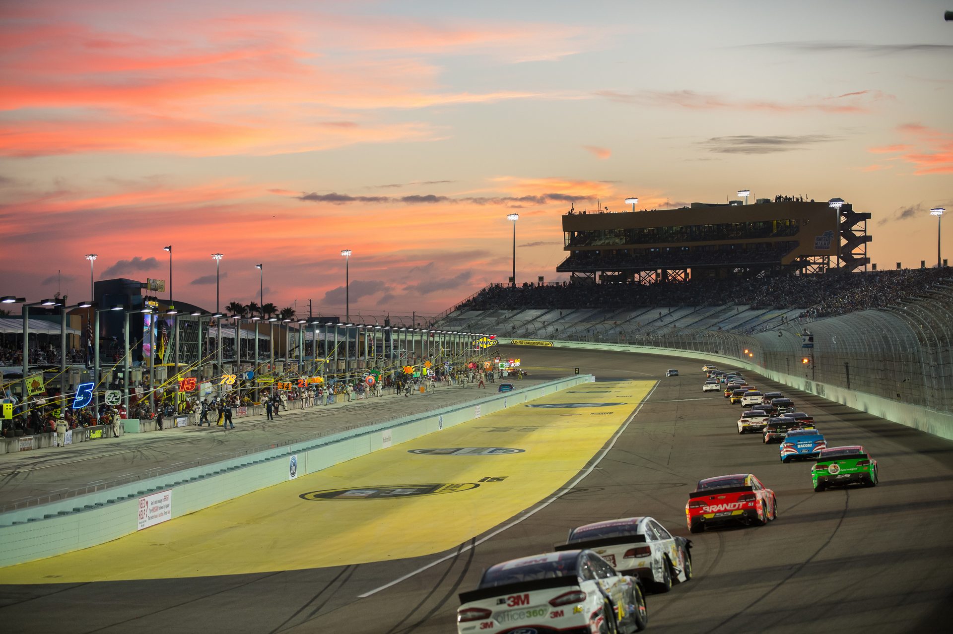 NASCAR stock cars race around a banked oval track at sunset, with pit row activity along the infield wall and packed grandstands silhouetted against a vivid pink and orange sky.