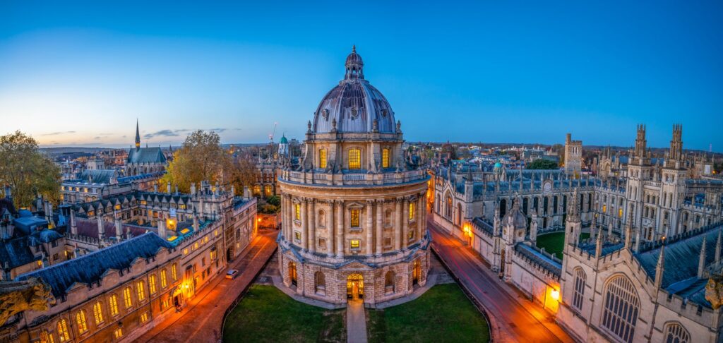 Aerial view of the Radcliffe Camera in Oxford, England, at dusk, with its iconic domed rotunda illuminated against a deep blue sky, surrounded by historic university buildings and glowing street lights.