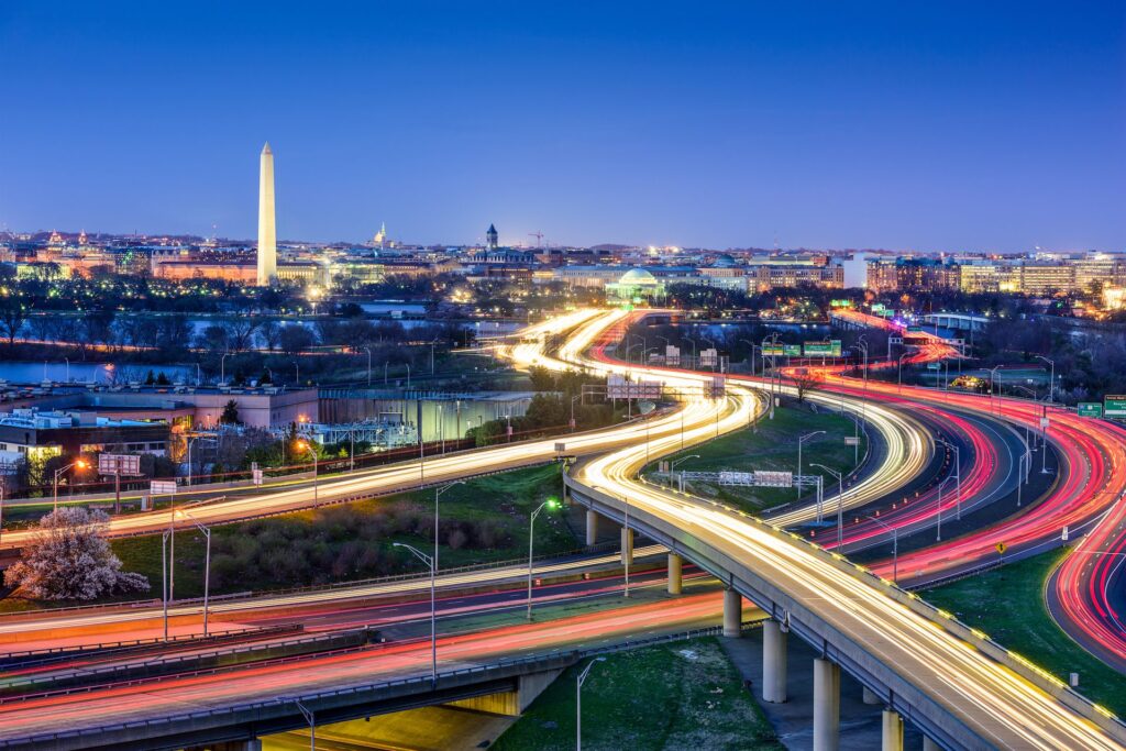 Long-exposure aerial view of Washington, D.C. at dusk, with streaking highway traffic light trails in the foreground and the Washington Monument and illuminated city skyline in the background.