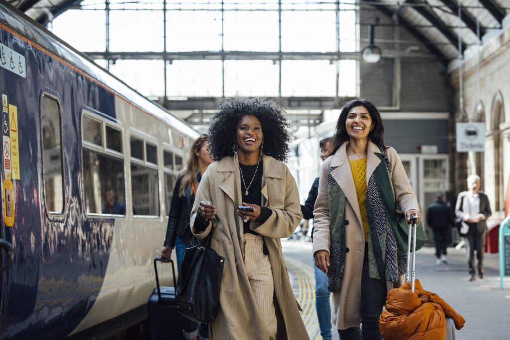 Two women smile and walk together along a train platform inside a historic station, each pulling luggage, with a passenger train alongside them.