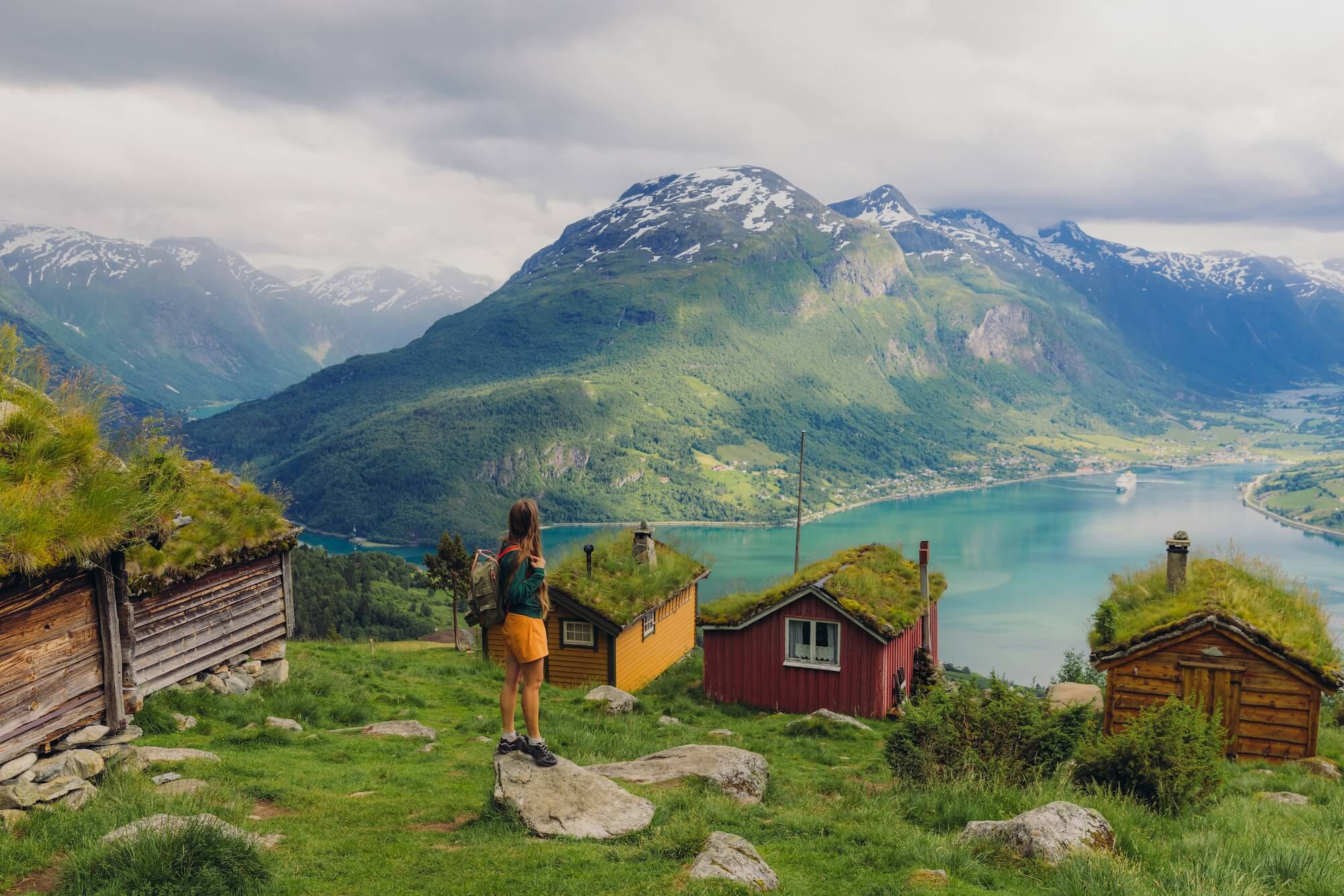 Hiker standing above a turquoise fjord beside grass-roofed cabins, with green mountains beyond.